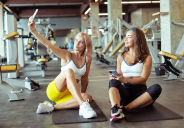 Spor salonundaki paspasın üzerinde otururken selfie çeken iki atletik bayan arkadaş.