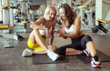 Spor salonundaki paspasın üzerinde dinlenirken akıllı telefon ekranına bakan iki gülümseyen atletik bayan arkadaş.