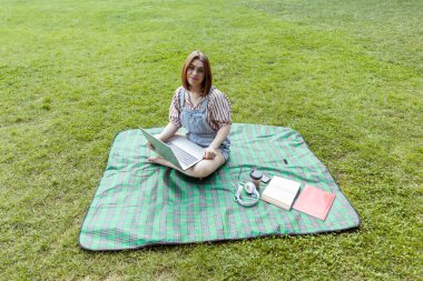 Parkta battaniyeye oturmuş bilgisayar kullanan genç bir kadın.