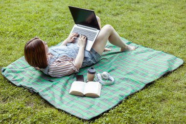 Genç kız öğrenci parkta bir battaniyenin üzerinde yatarken dizüstü bilgisayar kullanıyor.