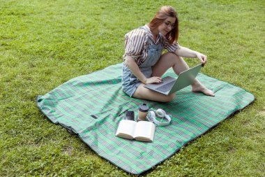 Parkta battaniyeye oturmuş bilgisayar kullanan genç bir kadın.