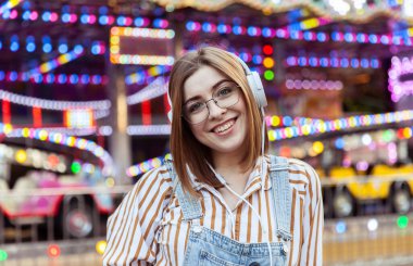 Kot tulumlu ve gözlüklü sevimli bir kadın lunaparkta kulaklıkla müzik dinliyor.