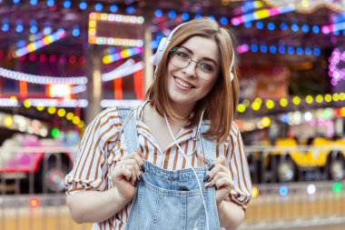 Kot tulumlu ve gözlüklü sevimli bir kadın lunaparkta kulaklıkla müzik dinliyor.