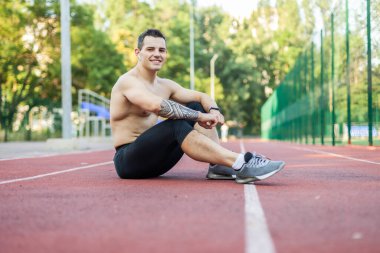 Stadyumda otururken çıplak gövdesiyle gülümseyen genç sporcu.