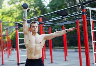 Çıplak gövdeli atletik bir erkek, spor sahasında tek bir dambılı var.
