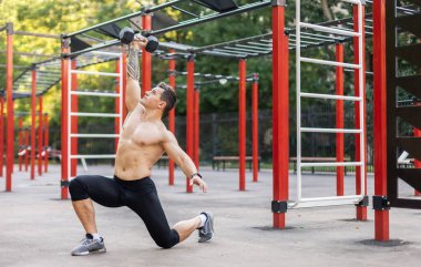 Çıplak gövdeli atletik bir erkek, spor sahasında tek bir dambılı var.
