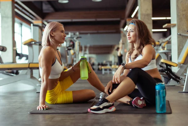 Spor salonunda paspasın üzerinde oturan iki atletik bayan arkadaş konuşuyor.