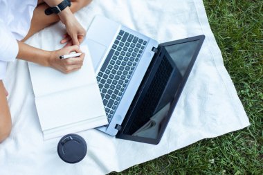Kadın, parkta bir dizüstü bilgisayarla battaniyeye otururken deftere yazıyor. Serbest çalışma, uzak çalışma veya eğitim