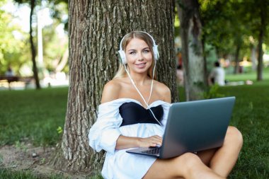 Kulaklıklı genç ve çekici bir kadın parkta çimenlere yaslanırken dizüstü bilgisayar kullanıyor. Serbest çalışma, uzaktan çalışma veya eğitim kavramı 
