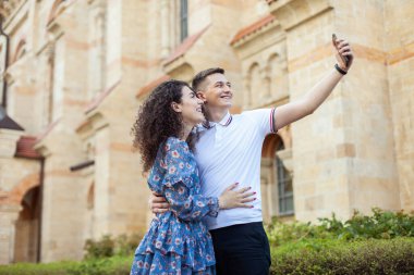 Young cheerful couple in love eating selfie on smartphone against urban architecture background