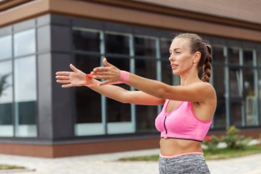 Formda bir kadın kol kaslarını dışarıda spor lastiği bantlarıyla çalıştırıyor.