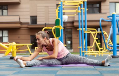 Dışarıdaki oyun parkında minderde otururken bacak esnetme alıştırması yapan formda bir kadın. Etkin yaşam biçimi