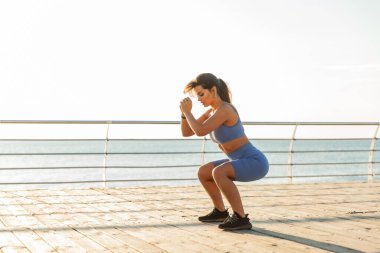 Güzel, kaslı, fit bir kadın sahilde çömelme egzersizi yapıyor.