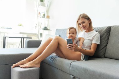 Sevimli genç bir anne ve küçük kızı evdeki kanepede otururken akıllı bir telefon kullanıyorlar.