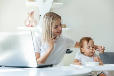 Work from home. Young mother is talking on phone while sitting at table with a laptop and feeding her little daughter. Motherhood, maternity, childhood and care concept.