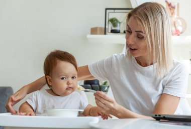 Şefkatli bir anne masada otururken bebeğini kaşıkla besler.