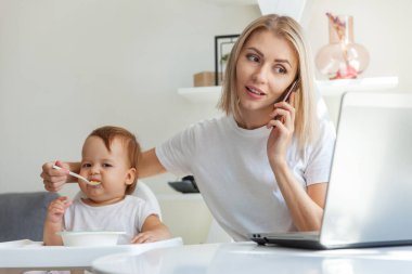 Evden çalışıyorum. İş anneleri telefonda konuşurken bir dizüstü bilgisayarla oturup küçük kızını besliyor. Annelik, annelik, çocukluk ve bakım kavramı.