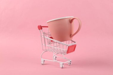 Pembe arka planda seramik bardaklı mini alışveriş arabası.