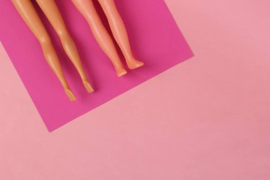 Plaj tatili kavramı. Oyuncak bebeklerin bacakları paspasın üzerinde uzanıyor ve pembe arka planda güneşleniyor. En küçük yerleşim