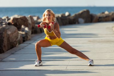 Sarı spor tişörtlü genç atletik kadın ve parlak güneşli bir günde kumsalda dambıllarla antrenman yapan kısa şortlu kadın.