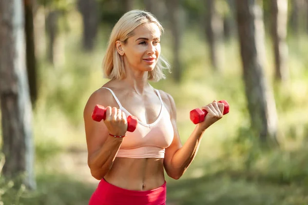 Young aesthetic woman in sportswear training with dumbbells in the forest