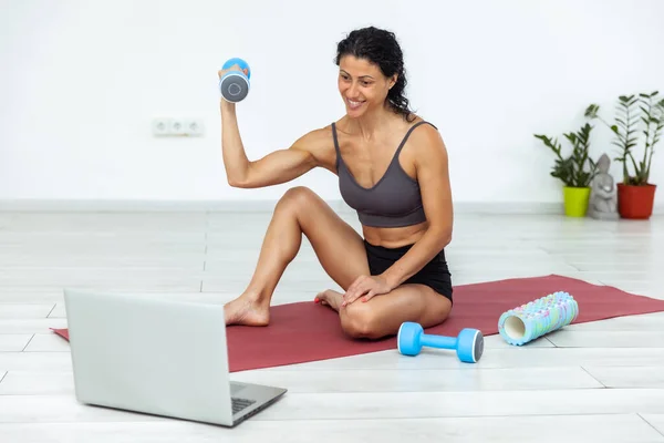 Online fitness class. Young athletic woman trainer exercising with dumbbell, broadcasting with laptop, recording her fitness blog or streaming indoors