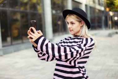 Genç, karizmatik, şapkalı bir kadın açık havada telefonda selfie çekiyor.