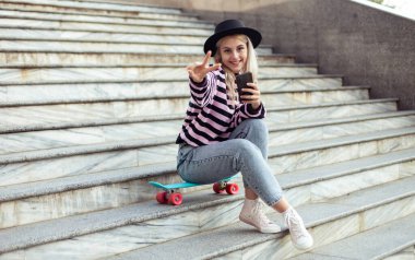 Şapkalı genç ve çekici bir kız merdivenlerde kaykayla otururken telefon kullanıyor.