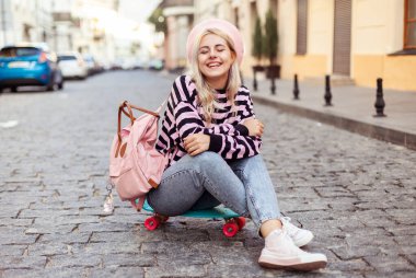 Sırt çantalı havalı kız Avrupa caddesinde kaykayın üzerinde oturuyor.