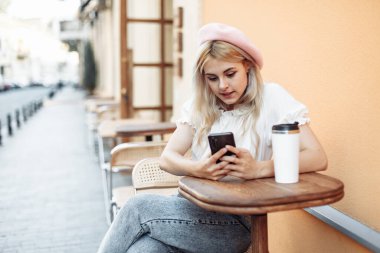 Bereli genç karizmatik kadın Avrupa caddesindeki açık kafede otururken telefon kullanıyor.