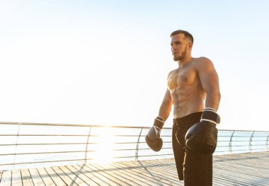 Sabahın erken saatlerinde çıplak gövdesi ve boks eldivenleriyle sahilde poz veren atletik bir adam.