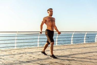 Young athletic man with a naked torso works out with fitness rubber bands in the early morning on the beach. Muscle training. Healthy lifestyle