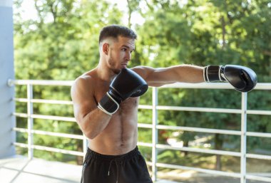 Açık havada boks eldivenli kaslı adam yumruklarını eğitiyor.