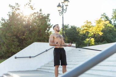 Çıplak gövdeli atletik kaslı adam dışarıda spor yaparken lastik bantlarla spor yapıyor.