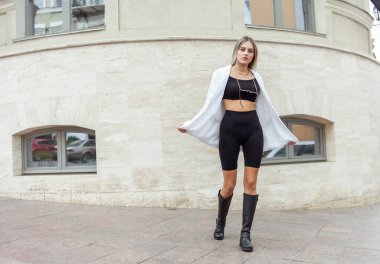 Beyaz ceketli, üst ve botlarıyla şehirde poz veren moda sarışın genç bir kadın. Sokak modası