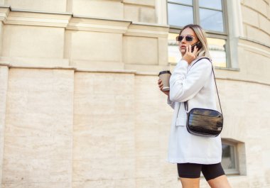 Moda kıyafetli genç sarışın kadın şehirde kahve içiyor ve telefonla konuşuyor.