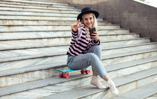 年轻迷人的嬉皮士女孩 戴着帽子 坐在楼梯上 户外滑板 一边用电话 — 图库照片