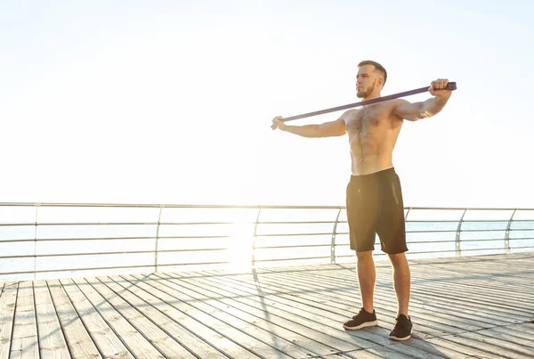 一个赤身裸体的年轻运动员清晨在海滩上与健身橡皮筋一起锻炼 肌肉训练 健康的生活方式 — 图库照片