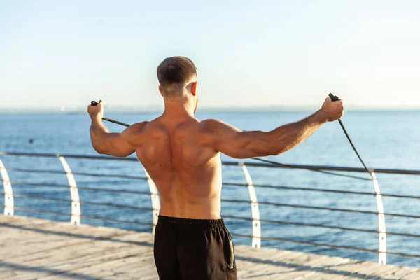 一个赤身裸体的年轻运动员清晨在海滩上与健身橡皮筋一起锻炼 肌肉训练 健康的生活方式 — 图库照片