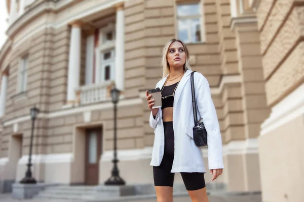 Young fashion blonde woman in trendy outfit drinks coffee on the go in city
