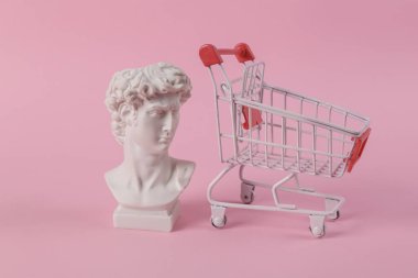 David 'in pembe arka planda alışveriş arabalı antika büstü. Kavramsal patlama. Minimum alışveriş hala hayattadır. Yaratıcı fikir