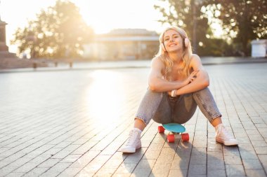 Genç, gülümseyen bir kadın şehirde kaykayın üzerinde otururken kulaklıkla müzik dinliyor.