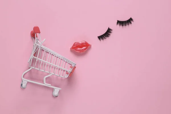 Beauty store. Shopping cart with lips and false eyelashes on pink background