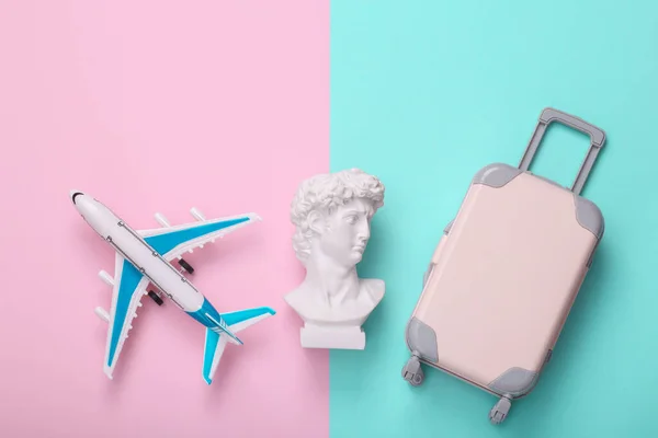 Toy baggage with air plane and David bust on pink blue background. Creative layout. Minimal travel still life. Flat lay. Top view