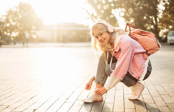 Sırt çantalı ve kulaklıklı sevimli bir kız öğrenci şehirde ayakkabı bağcıkları takıyor.