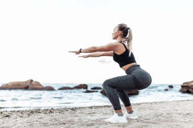 Beautiful young fit woman in sportswear practicing squats alone on wild beach. Healthy lifestyle concept