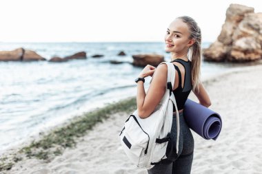 Fit woman with yoga mat and backpack posing alone on a wild beach
