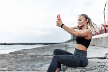 Fit woman in sportswear makes selfie on phone while sitting on urban beach
