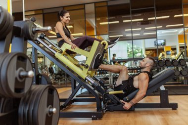 Strong man doing leg press in exercise machine to woman sitting on it in gym