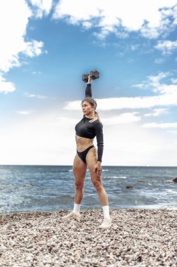 Strong athletic woman exercising with heavy dumbbell (duing overhead) on the beach during the day with blue sky and clouds. Functional outdoor training